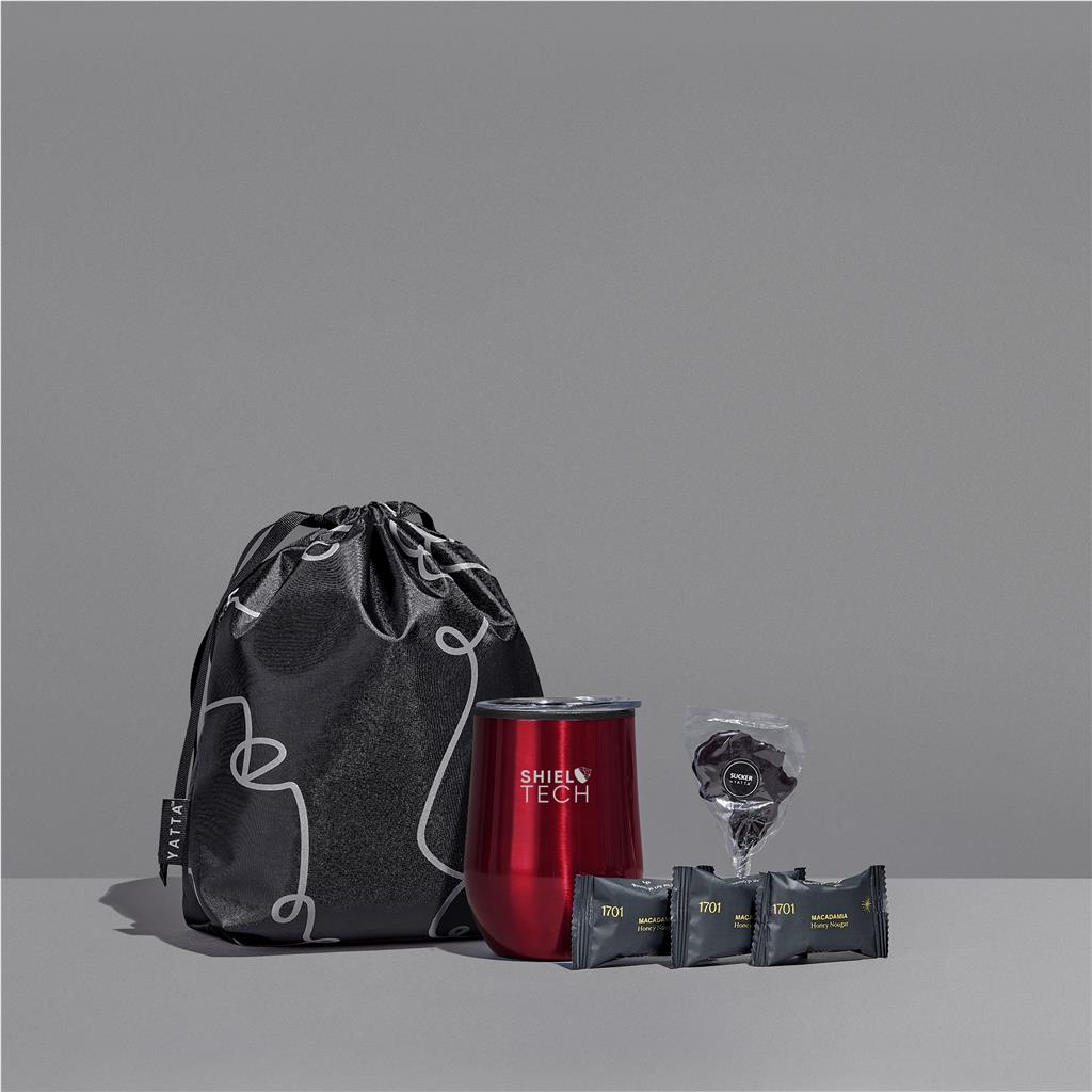 A product bundle featuring a black drawstring bag with white abstract line art, a metallic red insulated tumbler with 'SHIELD TECH' printed on the front, a small clear-lidded tumbler, and three dark snack packets, arranged on a gray surface against a gray background.