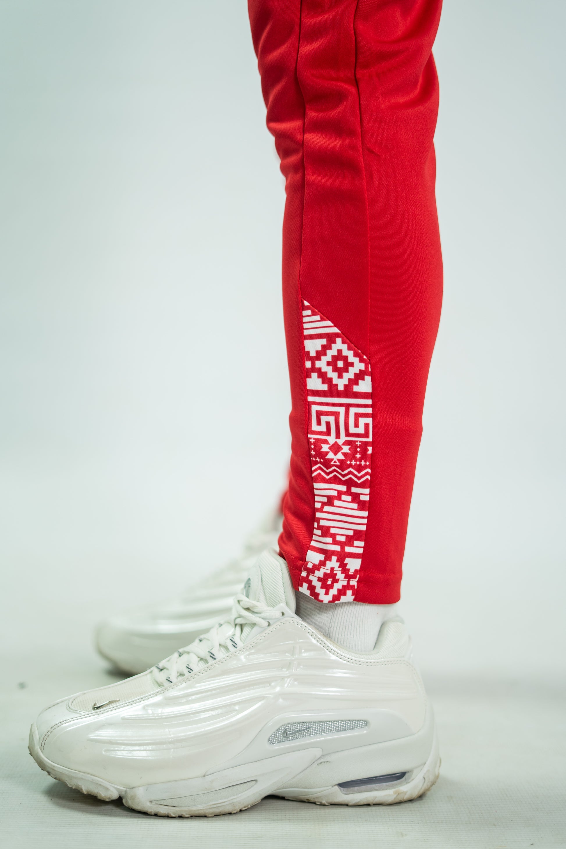 Red pants with a white geometric pattern on a leg, paired with white sneakers on a light gray background.

