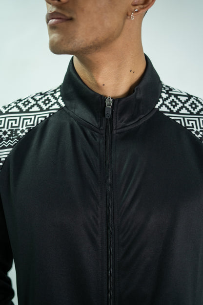 Person wearing a black jacket with a patterned shirt underneath on a plain background

