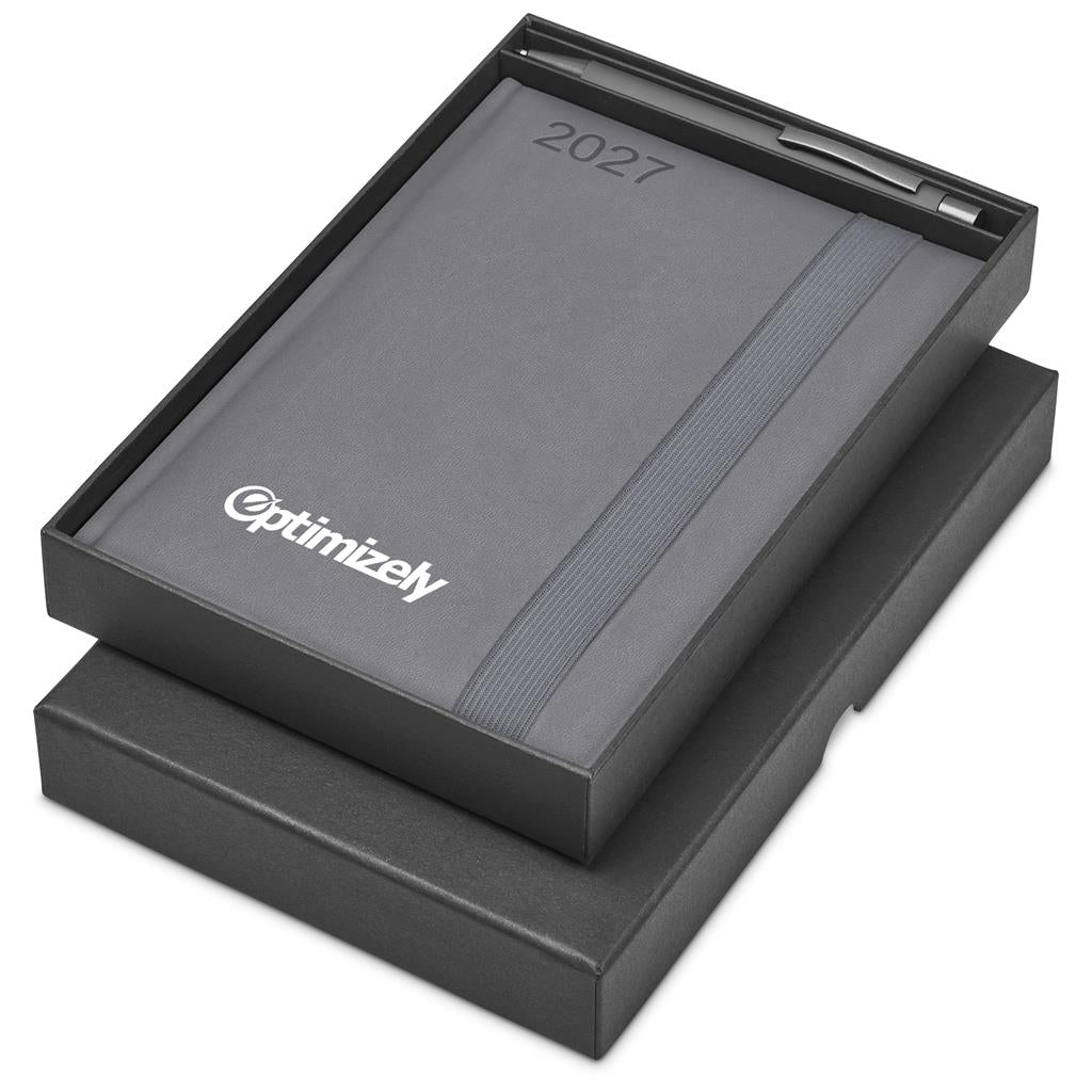 A 2027 Optimizely desk calendar in dark gray, shown open in its black presentation box with a pen inside, on a white background.

