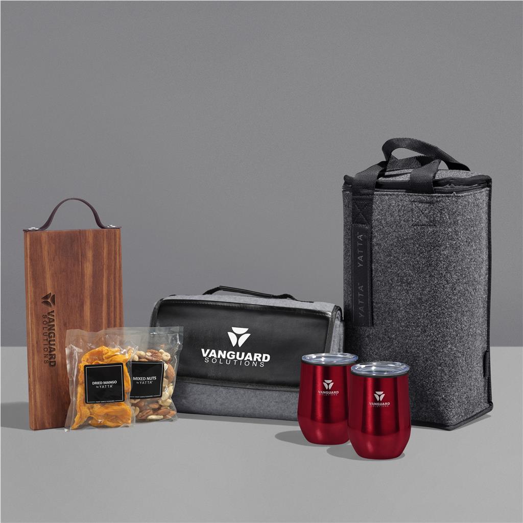 A branded gift set on a gray studio background, including a tall gray insulated tote bag, a black zippered pouch with white Vanguard Solutions branding, two red insulated tumblers with silver lids, a wooden box, and two clear bags of snacks.

