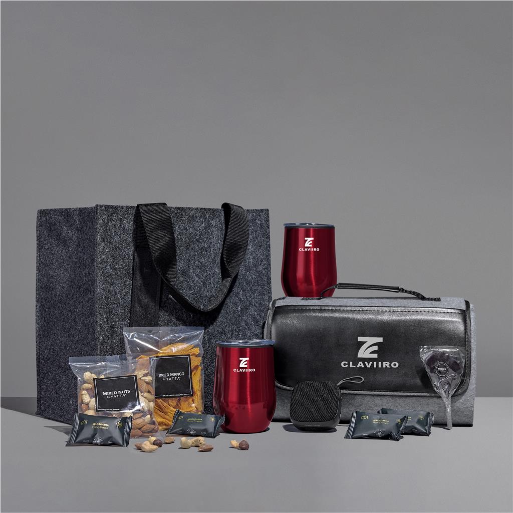 A Claviro-branded gift set arranged on a gray studio surface, including a dark gray felt tote bag, a metallic red insulated tumbler, a black-and-gray zippered case with Claviro branding, a small red tumbler, snack bags, nuts, and a glass.

