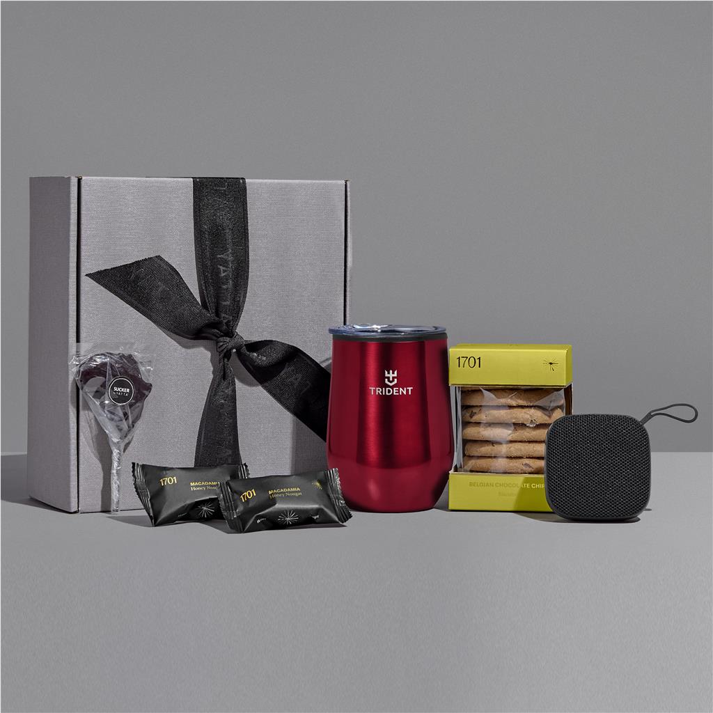 A gift set arranged on a gray surface against a gray background, including a large gray box with a black ribbon, a red Trident insulated tumbler, a yellow box of cookies, two black wrapped snacks, and a small black speaker.

