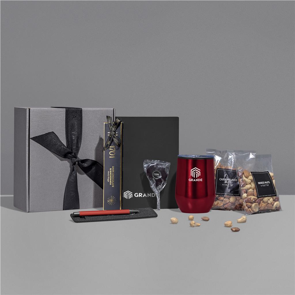 A gift set arranged on a light gray surface against a medium gray background, including a gray box with a black ribbon, a black branded package, a red insulated tumbler, a small clear bag, and two clear bags of nuts with black labels.

