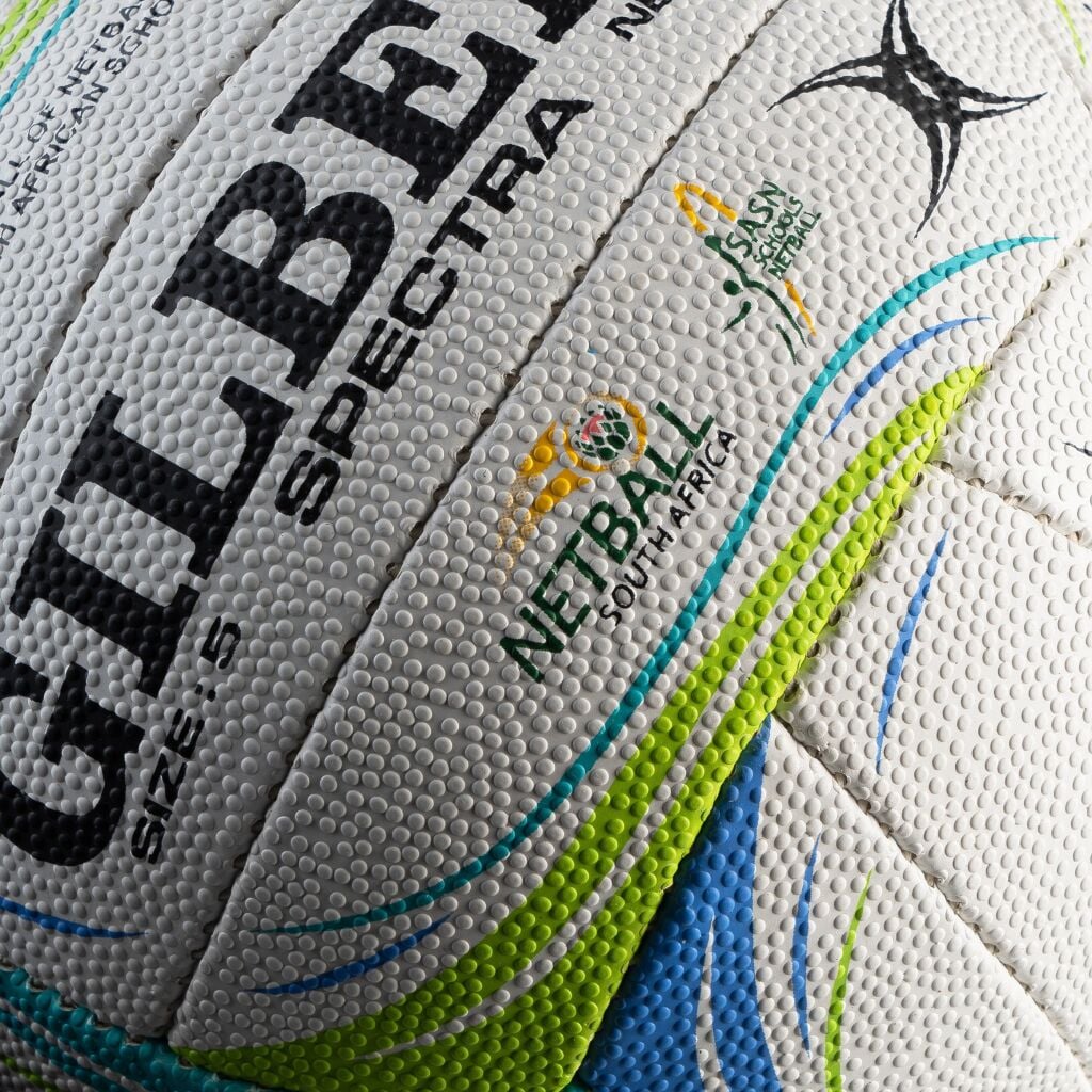 Close-up of a white textured rugby ball with black 'GILBERT' lettering, 'SPECTRA' text, and South Africa Netball branding with blue, green, and yellow accents.

