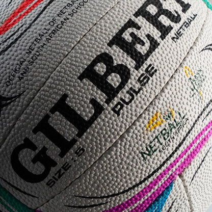 Close-up of a textured Gilbert netball ball showing the Gilbert brand name in black, with size 5 markings and South African Schools netball logos.


