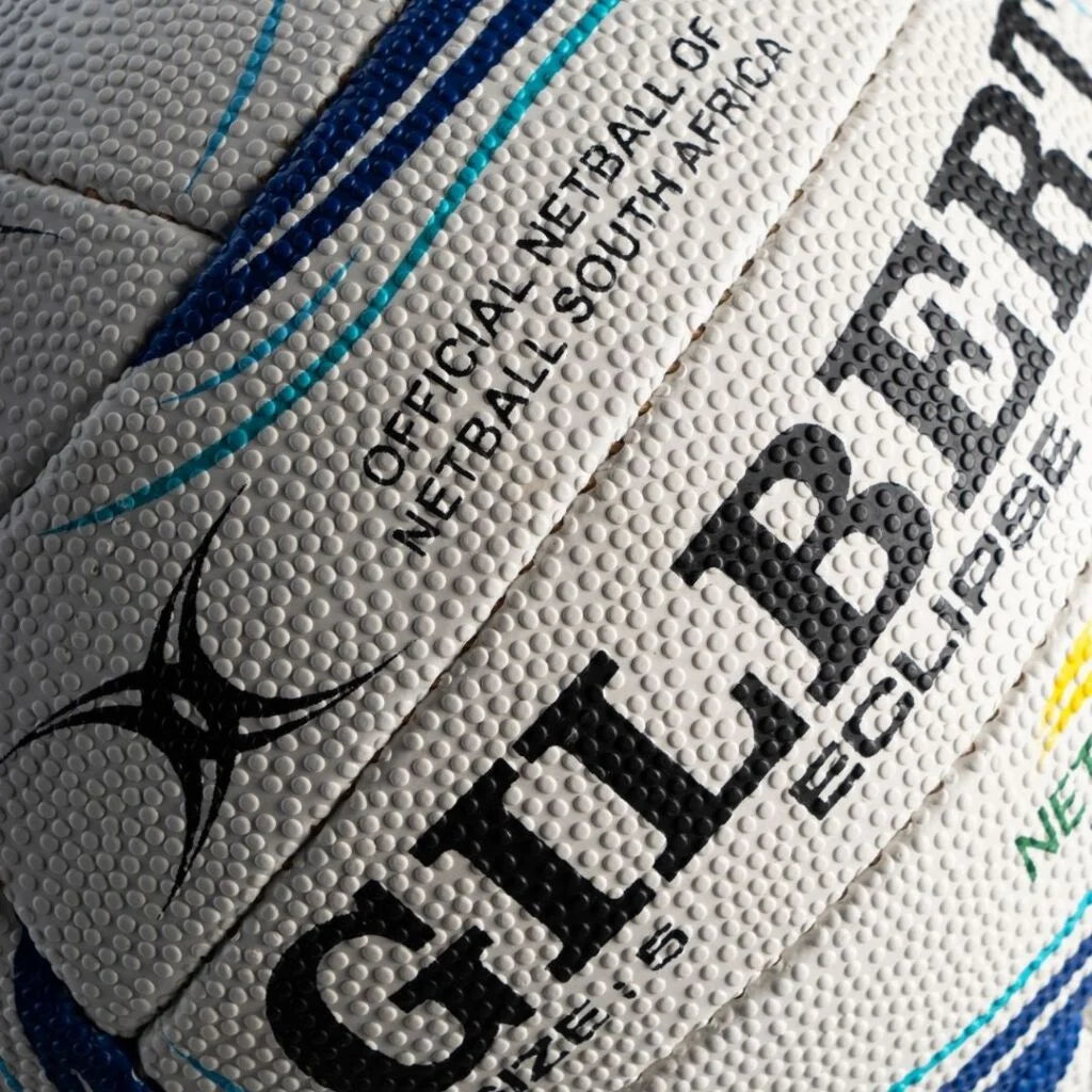 Close-up of a textured white netball with black and blue branding, including the Gilbert Eclipse logo and 'Official Netball of South Africa' text.

