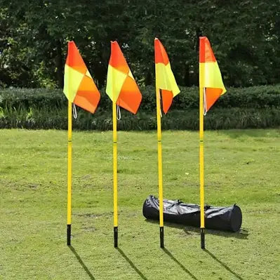 Four yellow and orange corner flags on yellow poles set in a grassy field, with a black carrying bag on the ground nearby and trees in the background.

